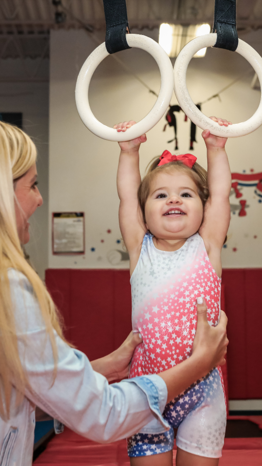 Toddler hanging from gymnastics rings with mom's help